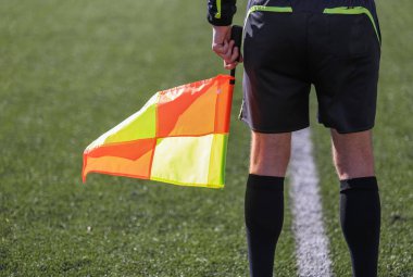Siyah takım elbiseli bir futbol hakemi elinde bayrakla bir futbol sahasında duruyor.