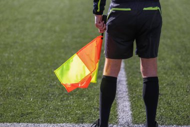 Siyah takım elbiseli bir futbol hakemi elinde bayrakla bir futbol sahasında duruyor.