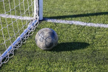 Bir futbol topu, parlak gündüz güneşinin altında bir futbol kalesinin yanındaki doğal yeşil çimlerin üzerinde yatar.