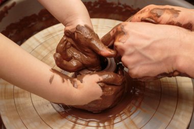 Çocuk çömlekçinin tekerindeki bir çömlek kalıbından şekilleniyor. Yetişkinler çocuğun kille çalışmasına yardım eder.