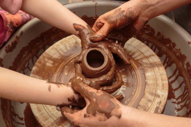 Çocuk çömlekçinin tekerindeki bir çömlek kalıbından şekilleniyor. Yetişkinler çocuğun kille çalışmasına yardım eder.