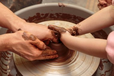 Çocuk çömlekçinin tekerindeki bir çömlek kalıbından şekilleniyor. Yetişkinler çocuğun kille çalışmasına yardım eder.