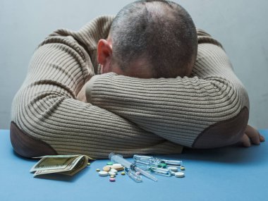 Masada bir adam var, yüzünü, parasını ve ilaçlarını saklıyor. Önündeki masada yatıyor..