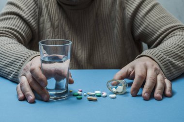 Masada bir adam, elinde bir bardak su, önüne saçılmış tıbbi haplar..