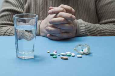 Masada bir adam, önünde bir bardak su ve etrafa saçılmış tıbbi haplar..