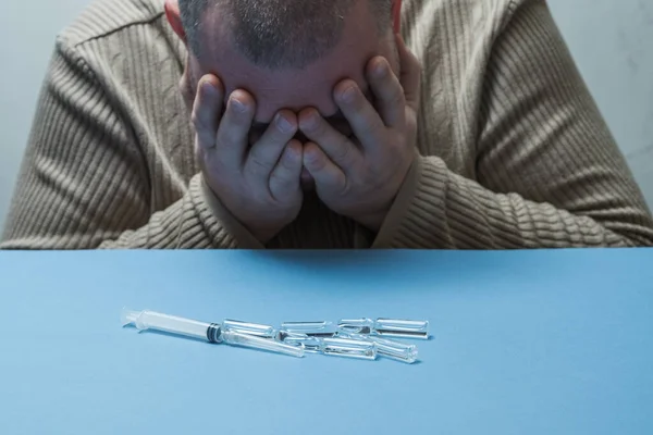 Adam elleriyle yüzünü gizliyor, şırıngalar ve ampuller önünde masanın üzerinde yatıyor..