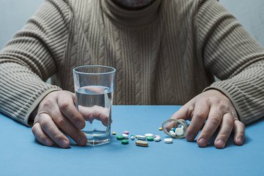 Masada bir adam, elinde bir bardak su, önüne saçılmış tıbbi haplar..