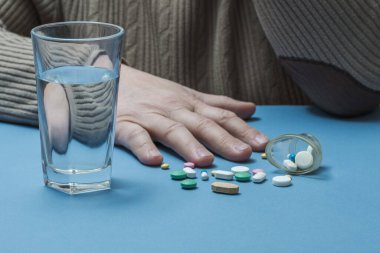 Masada bir adam, önünde bir bardak su ve etrafa saçılmış tıbbi haplar..