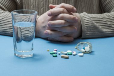 Önünde bir bardak su olan bir adam masada oturuyor ve sağlık hapları etrafa saçılıyor..