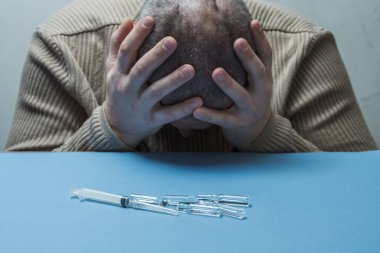 Bir adam masada oturuyor, elleriyle yüzünü gizliyor, şırıngalar ve ampuller önünde yatıyor..