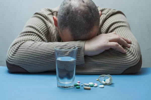 Adam elleriyle yüzünü gizler, jest belayı simgeler, ilaçlar ve bir bardak su önündedir..