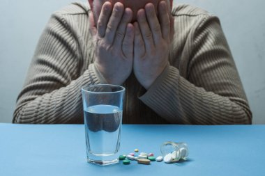 Adam elleriyle yüzünü gizler, jest belayı simgeler, ilaçlar ve bir bardak su önündedir..