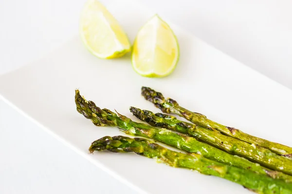 Asperges vertes cuites à l'huile d'olive et à l'ail. Servi avec deux morceaux de citron vert sur une assiette blanche . — Photo