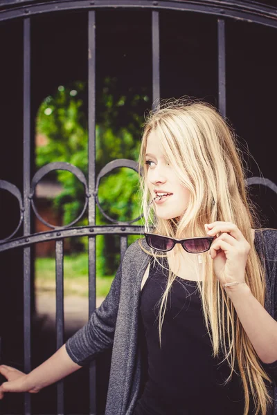 Belle fille blonde devant une vieille porte en fer. Beau jardin verdoyant dans la cour derrière la porte — Photo