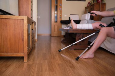 Olgun erkek ve yaralı bacağı televizyon izlerken uzaktan kumanda kullanıyor.