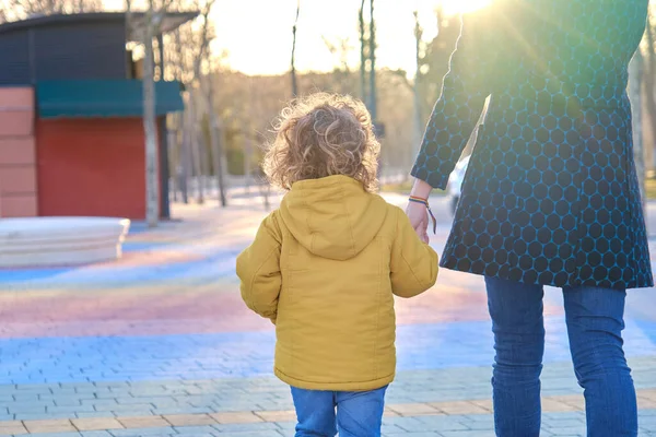 Una madre caminando de la mano con su hijo pequeño disfrutando de un momento de convivencia familiar en un parque al atardecer. — Foto de Stock