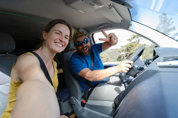 Positive man showing peace sign and woman looking at camera while taking self portrait in automobile during road trip in countryside on summer day