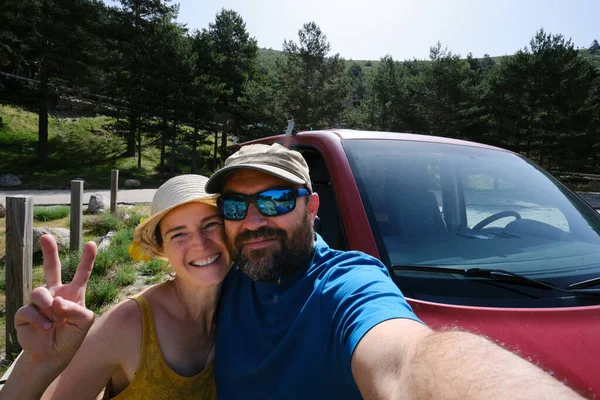 Delighted woman with peace sign and man taking self portrait while standing near automobile during road trip in nature on summer day