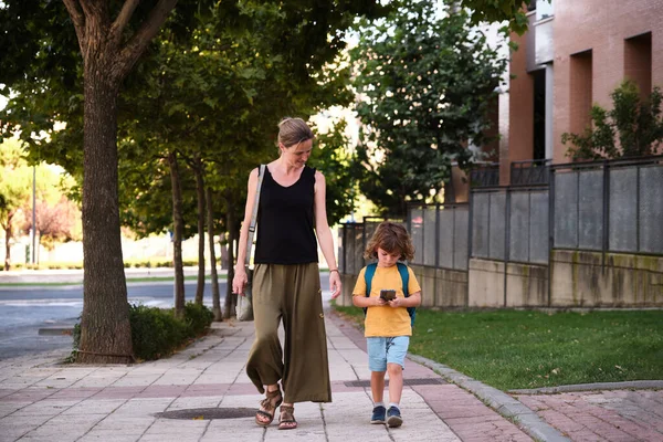 Mother and son walking down the street after school. The child is using the smart phone