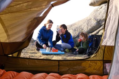 Children sipping hot drink and eating apple while sitting cross legged near mother and having breakfast outside tent in mountains
