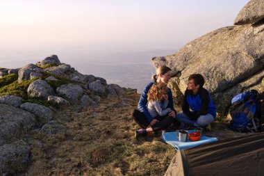 Mom embracing child and talking with son while sitting cross legged on ground near stone at sunrise on campsite