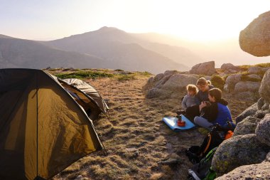 Mom with kids sitting on ground near tent and enjoying hot drink early in morning in highlands