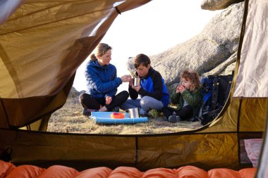 Children sipping hot drink and eating apple while sitting cross legged near mother and having breakfast outside tent in mountains