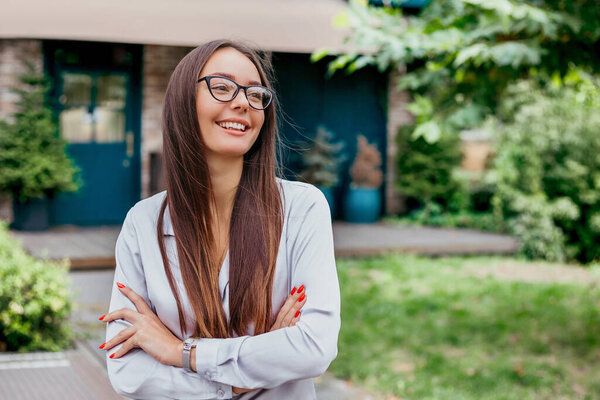 Молодая студентка в очках улыбается со сложенными руками, одетыми в рубашку, на фоне фасада здания, копирует пространство. Крупный план портрета молодой женщины в очках