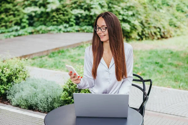 A young brunette woman in glasses sits at a table in a cafe and works at a laptop. Remote work, freelance, job search. Blogging. Online training