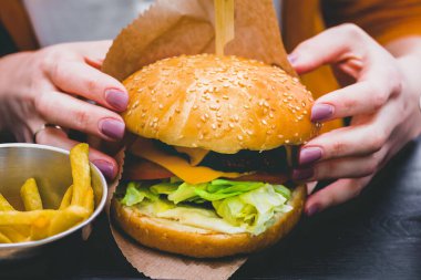 Siyah ahşap masada patates kızartması ve taze lezzetli bir burger tutan kadın eller.