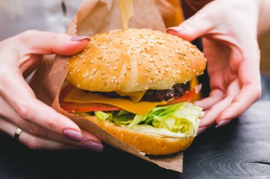 Siyah ahşap masada patates kızartması ve taze lezzetli bir burger tutan kadın eller.