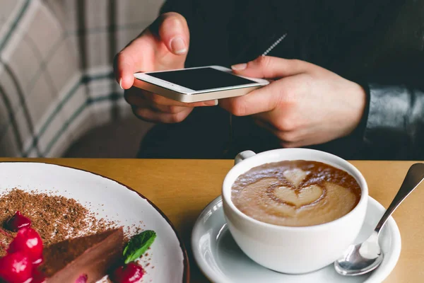 Kafede cep telefonu, kahve ve kek olan bir kız. Akıllı telefon tutan bir kadın eli. Kız bir kafede sohbet ediyor ya da internetten alışveriş yapıyor, sosyal medyayı kontrol ediyor ya da cep telefonuyla sohbet ediyor. Kahve kapuçinosu çiziyorum.