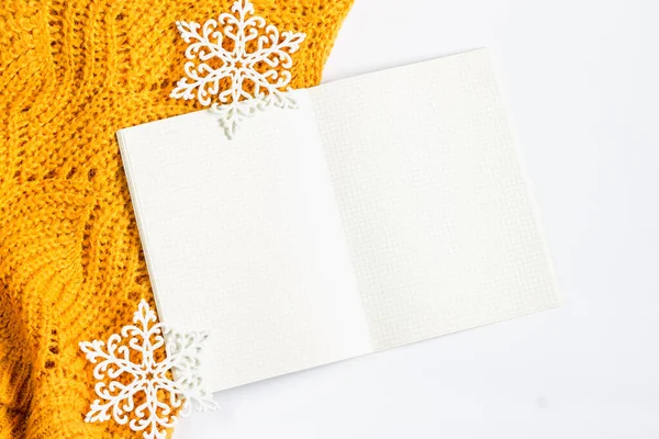 Open notepad with sheets, orange sweater, decorative snowflakes on a white background, flat lay, top view, copy space for text