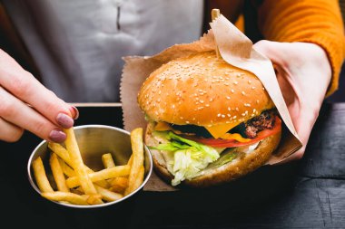 Siyah ahşap masada patates kızartması ve taze lezzetli bir burger tutan kadın eller.