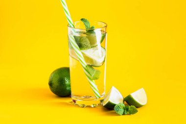 Suyla taze yaz limonatası, limon dilimleri ve sarı arka planda pipetle cam bir bardak içinde nane şekeri. Soğuk, ferahlatıcı yaz kokteyli. Kopya alanı ile taze içecek kompozisyonu