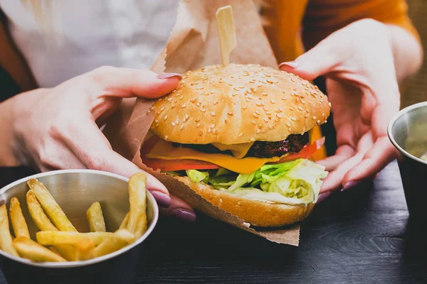 Siyah ahşap masada patates kızartması ve taze lezzetli bir burger tutan kadın eller.