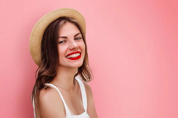 A young girl in straw hat and red lipstick smiling on a pink background, copy space. Tourist girl having fun. Happy girl looking at the camera smiling and laughing, space for text