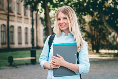Sarışın bir öğrenci kız gülümsüyor ve üniversite arka planında elinde bir klasör ve defter tutuyor. Eski bir üniversitenin arka planında bir kız duruyor..