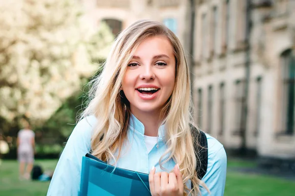 A blonde student girl is laughing and holding a folder and a notebook in her hands at university background. A girl stands on the background of an ancient university, copy space