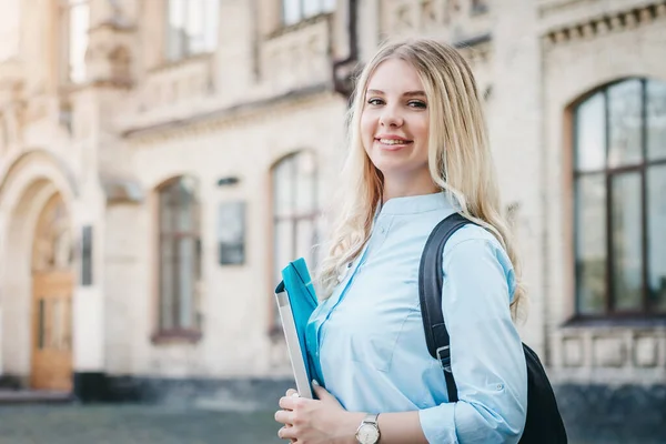 A blonde student girl is smiling and holding a folder and a notebook in her hands on a university background. Girl is taking exams at university