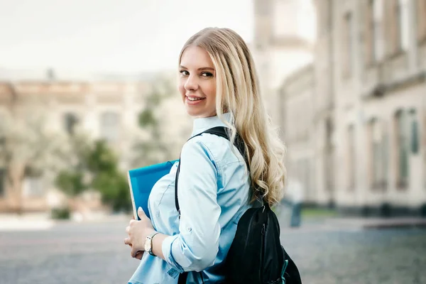 一个金发碧眼的女学生笑着 手里拿着一个文件夹和一个笔记本 手里拿着一本大学背景的书 女孩在大学里参加考试 — 图库照片