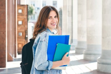 Üniversiteye karşı gülümseyen genç bir kız. Sevimli kız öğrenci klasörleri ve defterleri elinde tutuyor. Öğrenme, eğitim kavramı