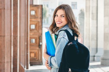 Üniversiteye karşı gülümseyen genç bir kız. Sevimli kız öğrenci klasörleri ve defterleri elinde tutuyor. Öğrenme, eğitim kavramı