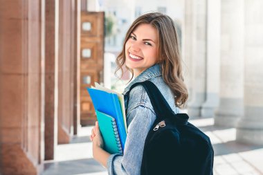 Üniversiteye karşı gülümseyen genç bir kız. Sevimli kız öğrenci klasörleri ve defterleri elinde tutuyor. Öğrenme, eğitim kavramı