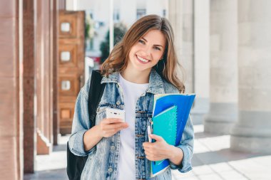 Üniversiteye karşı gülümseyen genç bir kız. Sevimli kız öğrenci klasörleri, defterleri ve cep telefonunu elinde tutuyor. Öğrenme, eğitim kavramı