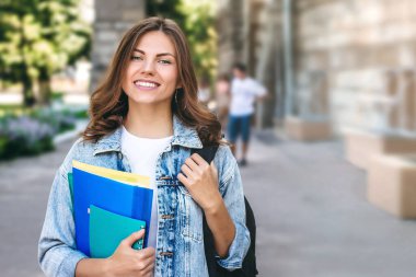 Üniversiteye karşı gülümseyen genç bir kız. Sevimli kız öğrenci klasörleri ve defterleri elinde tutuyor. Öğrenme, eğitim kavramı