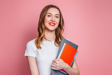 Eğitim konsepti. Mutlu beyaz öğrenci kız gülümsüyor. Kameraya bakıyor ve elinde defterler var. Pembe stüdyo arka planında izole edilmiş kitaplar.