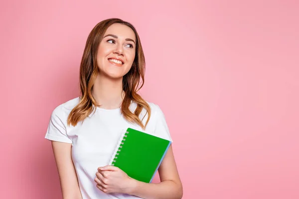 student girl smiles and stands isolated over pink background and look away.copy space. Portrait of a young woman holding a notepad, notebooks for study. Girl learning english lessons