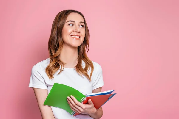 student girl smiles and stands isolated over pink background and look away. Portrait of a young woman holding a notepad, notebooks for study. Girl learning english lessons
