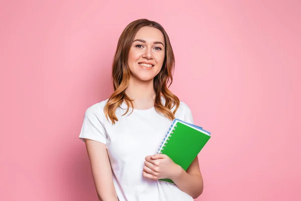 student girl smiles and stands isolated over pink background. Portrait of a young woman holding a notepad, notebooks for study. Girl learning english lessons